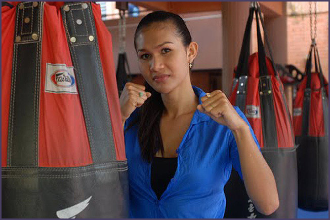 Parinya Charoenphol poses with fists raised in front of a red Fairtex punching bag, wearing a bright blue button-up shirt with a black top underneath, inside a Muay Thai gym.